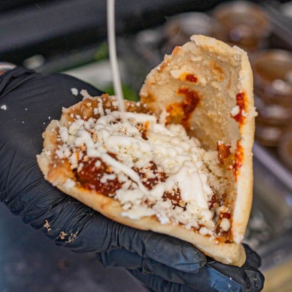 A hand in a black glove holds a Mexican gordita filled with red sauce, cheese, and crema.
