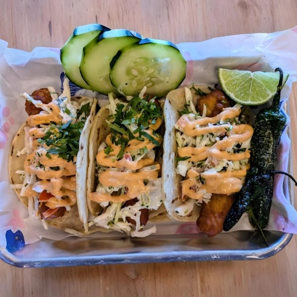 Three fish tacos with cabbage, cilantro, and orange sauce, served with cucumber, lime, and a pepper.