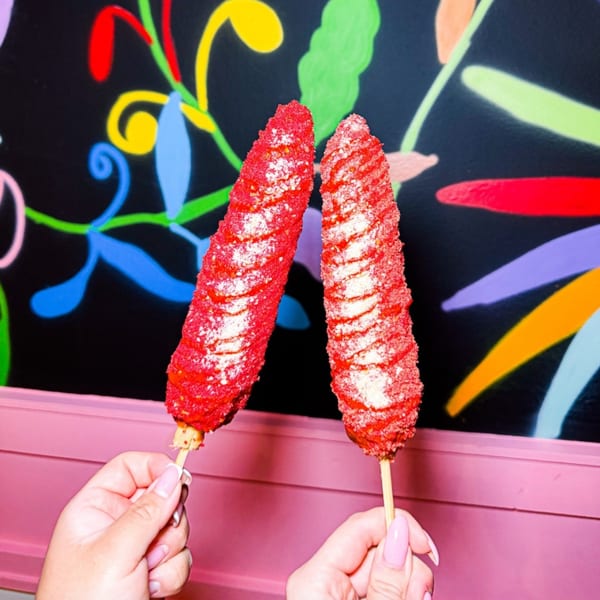 Two hands holding red elotes with white cheese against a colorful mural at Fonda Lupita.
